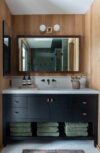 Modern mountain homes bathroom vanity with dark cabinetry, open towel storage, wall-mounted faucet, mirror, and wood wall paneling