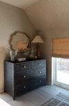 Modern mountain homes bedroom detail with dark wood dresser, scalloped mirror, table lamp, roman shade, and soft natural light