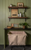 Modern mountain homes laundry room detail with green paneled wall, open wood shelving, black counter, and canvas laundry bin