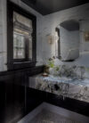 Coastal interiors bathroom vanity with dark cabinetry, marble counter, arched mirror, window shade, and wall sconces