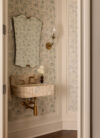 Bathroom sink area with wall-mounted marble basin, patterned wallcovering, scalloped mirror, sconces, and brass-toned fixtures