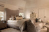 Luxury whole house renovation NJ bedroom with upholstered bed, layered neutral bedding, accent chair, bench seating, dark wood nightstands, and short windows