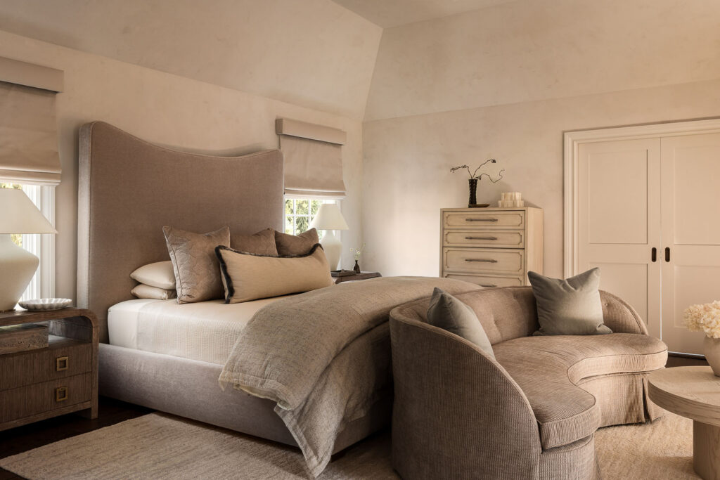 Luxury whole house renovation NJ bedroom with upholstered bed, layered neutral bedding, accent chair, bench seating, dark wood nightstands, and short windows