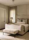 Bedroom with upholstered bed, scalloped headboard, bench seating, tall drapery, textured wall, and bedside lamp atop a wood night stand