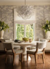 Luxury whole house renovation NJ dining nook with round table, upholstered chairs, patterned wallcovering, large window, sculptural chandelier, and tall greenery