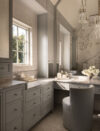 Vanity and dressing area with curved upholstered stool, built-in drawers, tall mirror, window, and chandelier