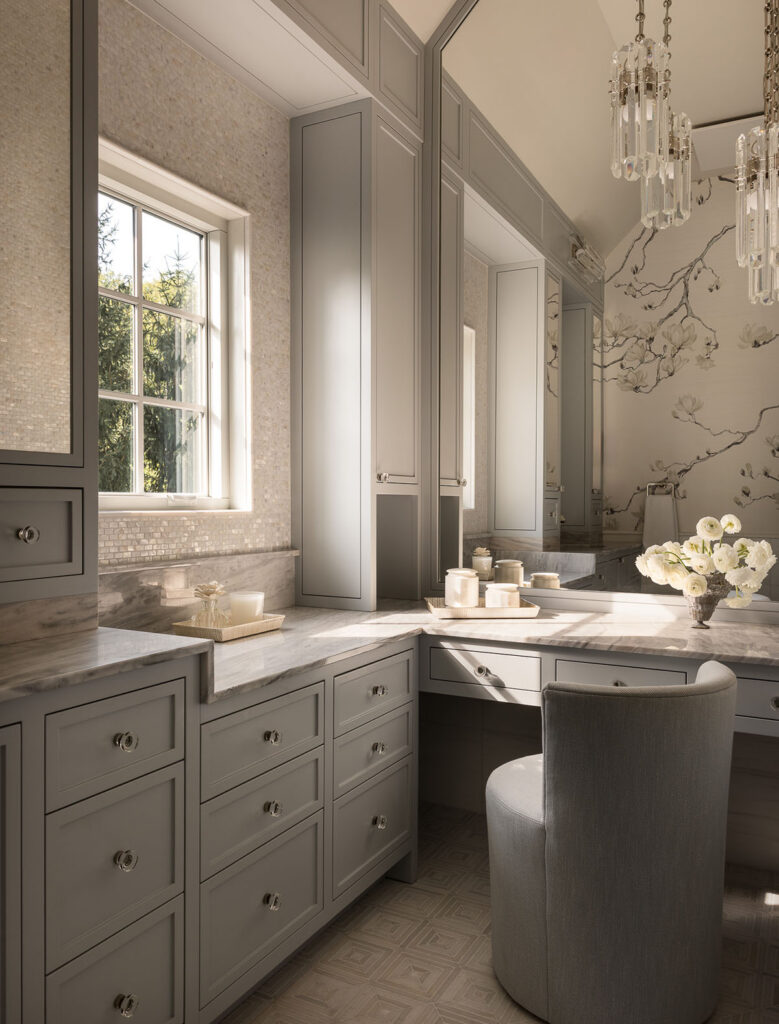 Vanity and dressing area with curved upholstered stool, built-in drawers, tall mirror, window, and chandelier