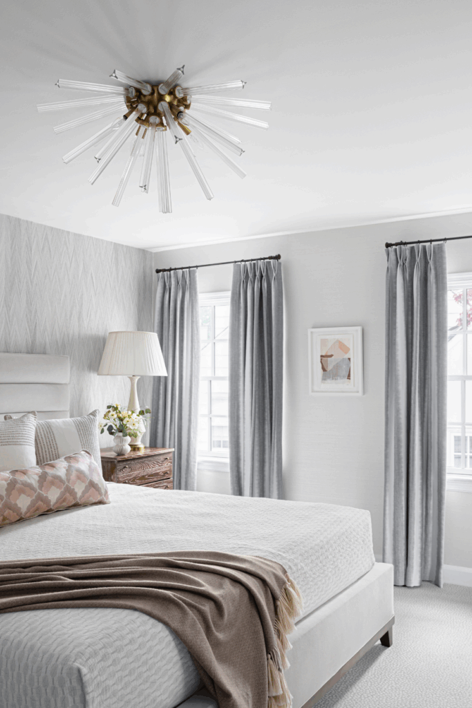 Bright bedroom with upholstered bed, layered neutral bedding, gray curtains, and modern ceiling light fixture