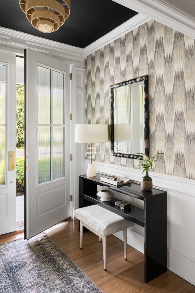 Modern entryway with black console table, patterned wallpaper, upholstered bench, mirror, and layered lighting